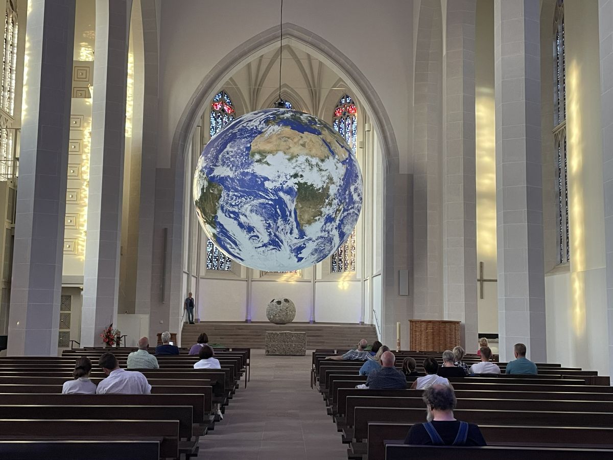 In einem Kirchenraum mit Kirchenbänken und Besucher*innen hängt eine sehr große Weltkugel.