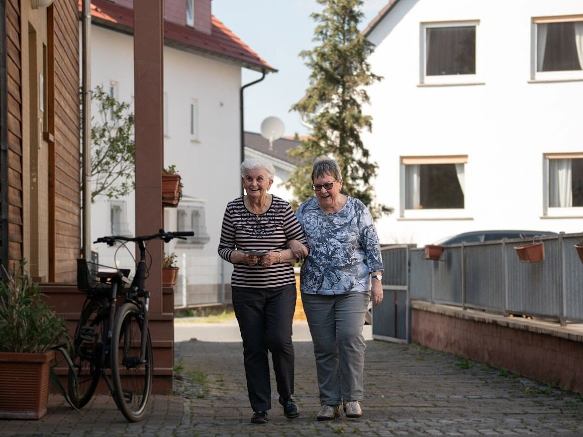 Zwei ältere Frauen gehen eingehakt spazieren vor einem Wohnhaus.