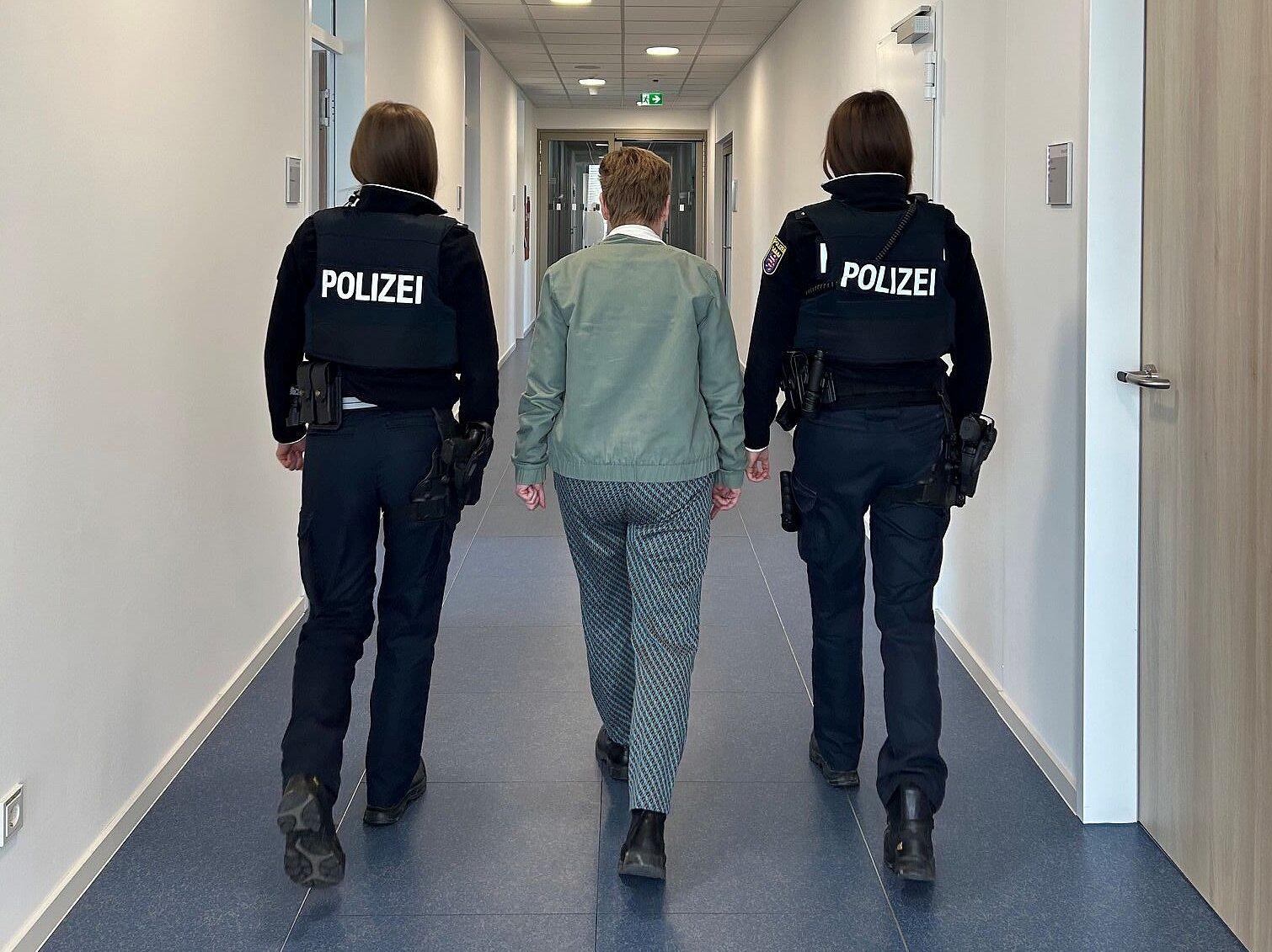 Zwei Polizistinnen in Uniform gehen einen Bürogang entlang zwischen ihnen geht eine Frau mit grüner Jacke. 