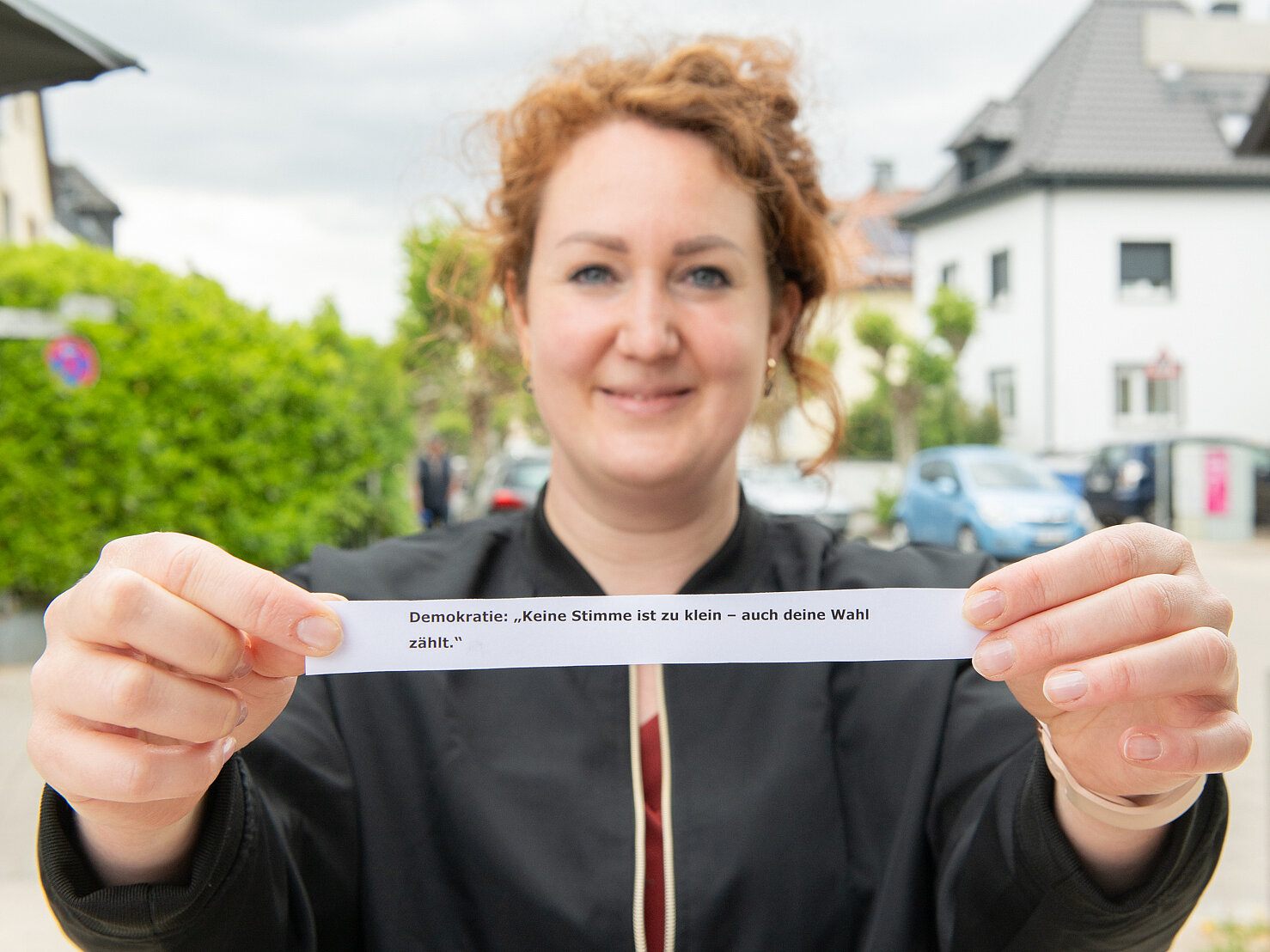Eine Frau mit schwarzer Jacke und braunen Locken streckt eine Zettel engegen auf dem steht: Demokratie: keine Stimme ist zu klein - auch deine Wahl zählt.