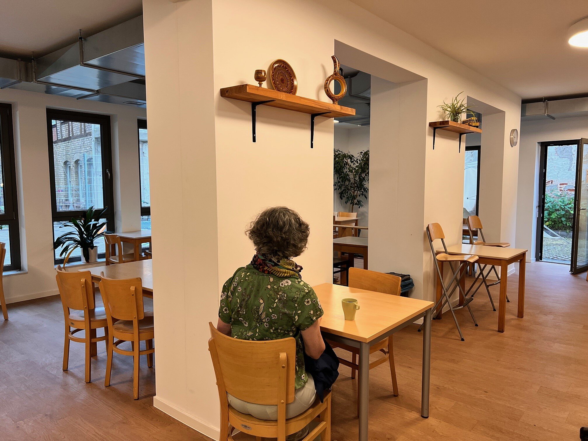 In einem Raum mit Holzboden und Holzstühlen sitzt eine Frau, man sieht sie von hinten, vor ihr steht eine Tasse Kaffee.e