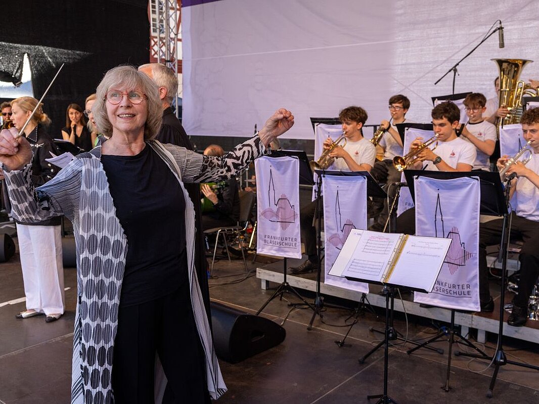 Eine Frau mit mit grauer gemusterter Jacke steht vor einem Orchester von Jugendlichen und dirigiert