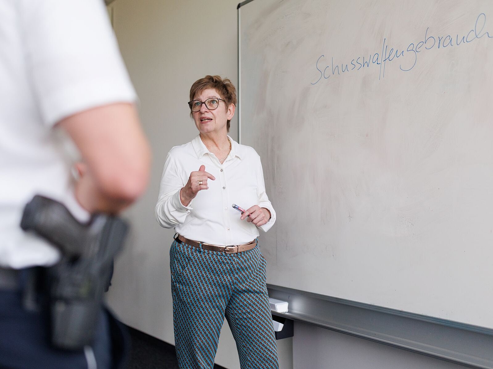 Eine Frau mit weißer Bluse steht an einer Tafel, ein Mensch steht vor ihr mit einer Schusswaffe im Halfter.