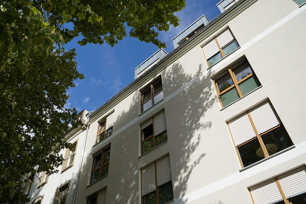 Eine weiße Hauswand von unten nach oben fotografiert mit vielen Fenstern, dahinter blauer Himmel.