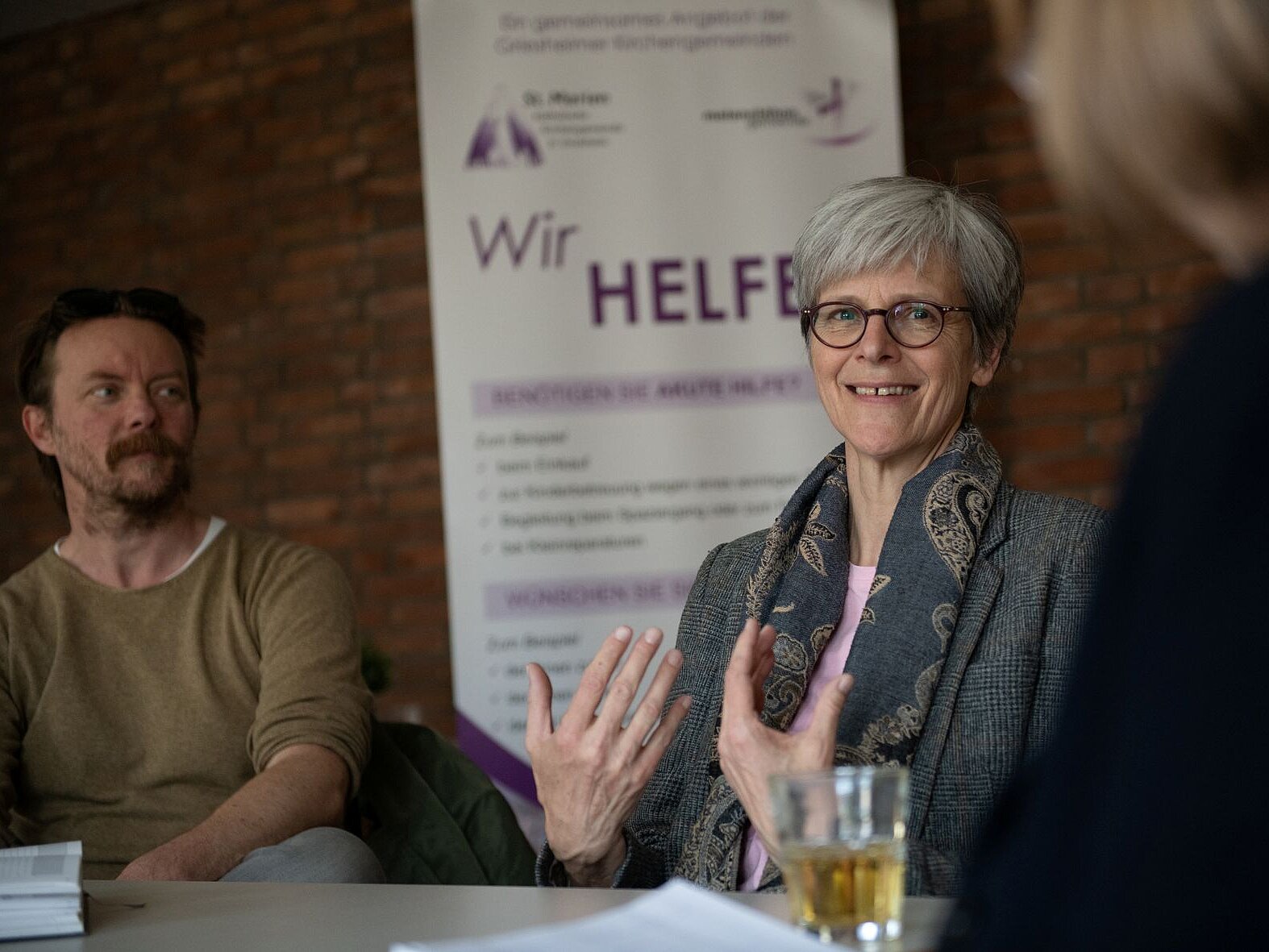 Eine Frau mit grauen Haaren sitzt zwischen zwei Menschen am Tisch und spricht, dabei gestikuliert sie mit ihren Händen. 