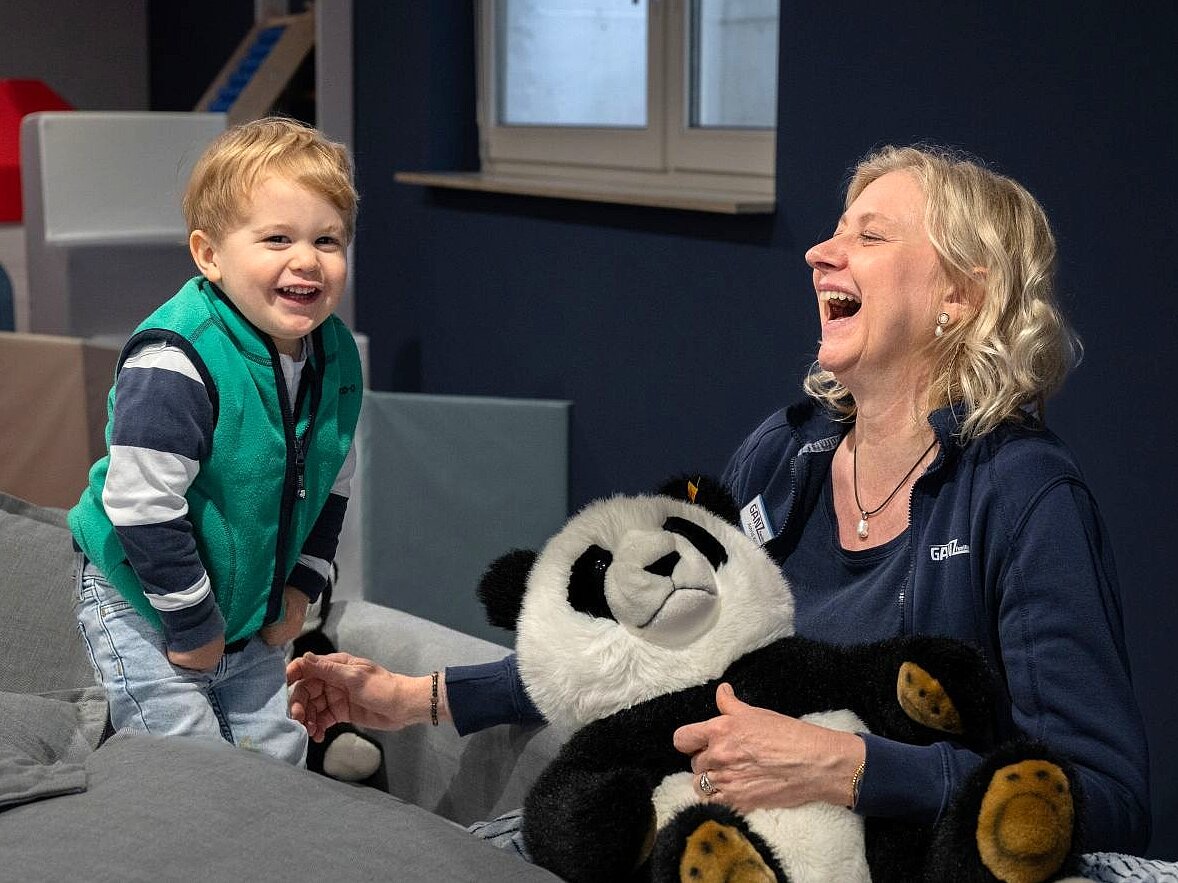 Eine Frau und ein kleiner Junge sitzen sich in einer Wohnung lachend gegenüber, die Frau hält einen Plüsch-Pandabären in der Hand.